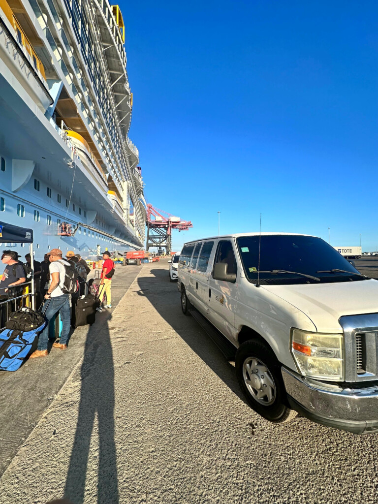 People with luggage waiting beside a white shuttle van and cruise ship at the San Juan port.